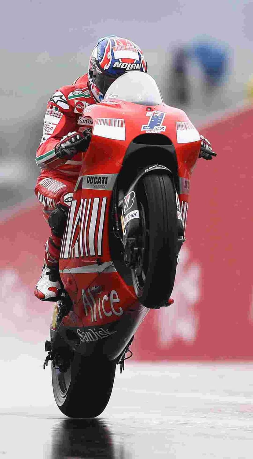 MotoGP racer on a red Ducati performs a high-speed wheelie on a wet asphalt track during a professional race.