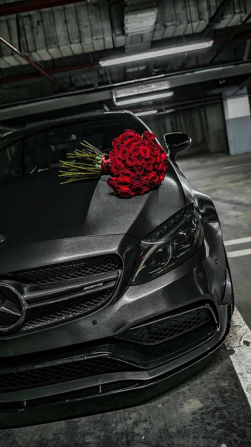 Dark grey luxury sedan with a large bouquet of red roses on the hood inside a dimly lit garage setting.