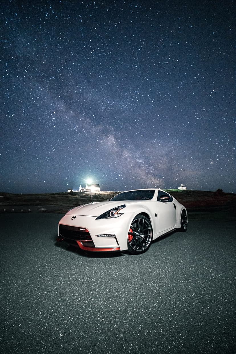 White Nissan 370Z sports car parked on an asphalt road at night under a star-filled Milky Way galaxy sky.