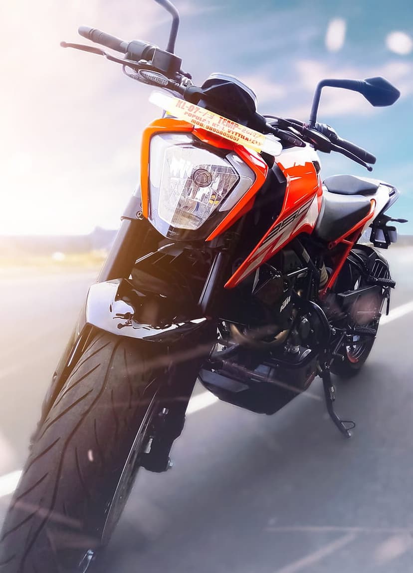 KTM Duke motorcycle front view with a sharp headlight, orange frame, and lens flare on a sunlit asphalt road.