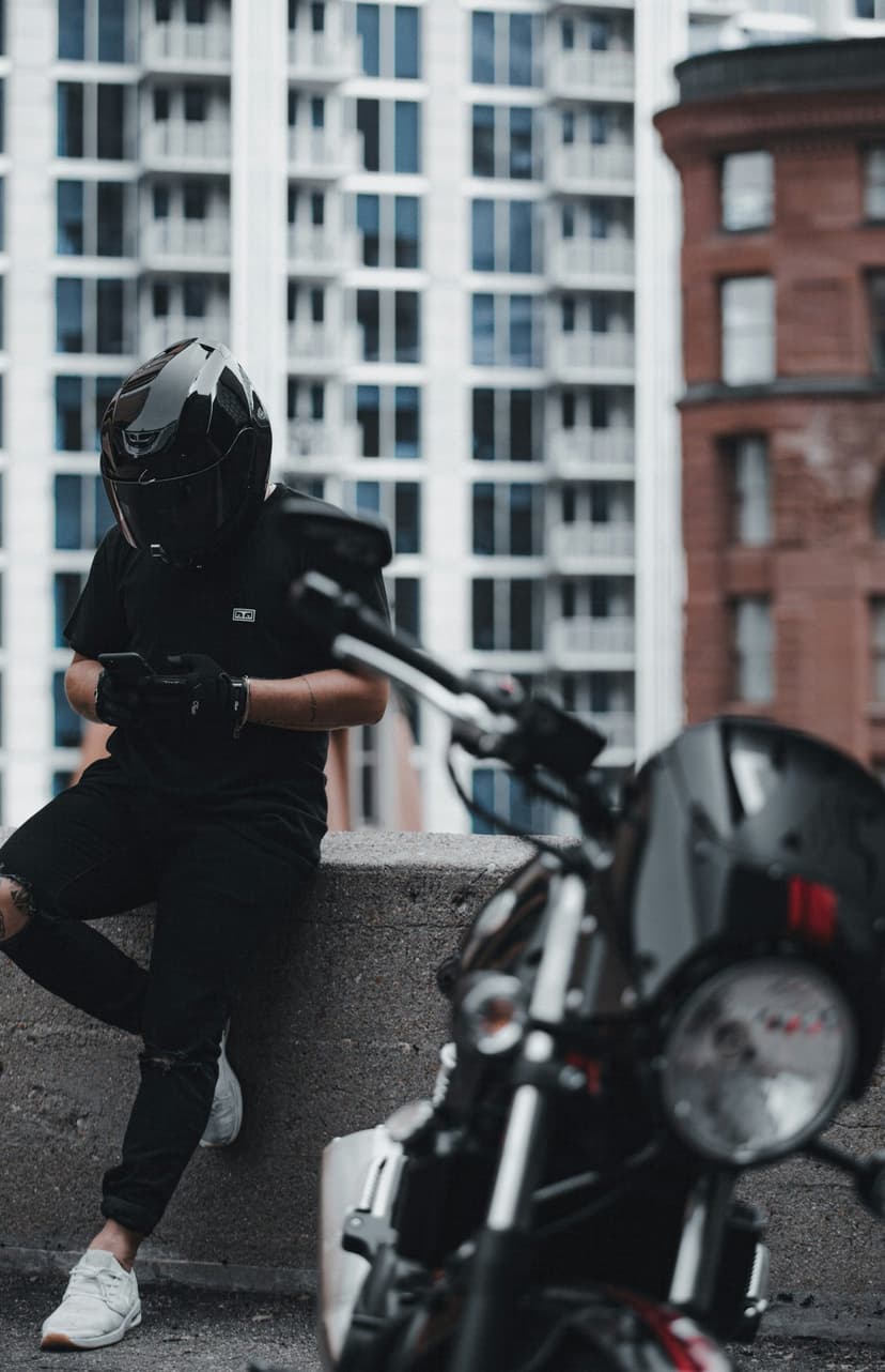 Motorcyclist in a black helmet sits on a stone ledge checking a phone with a blurred bike in the foreground.