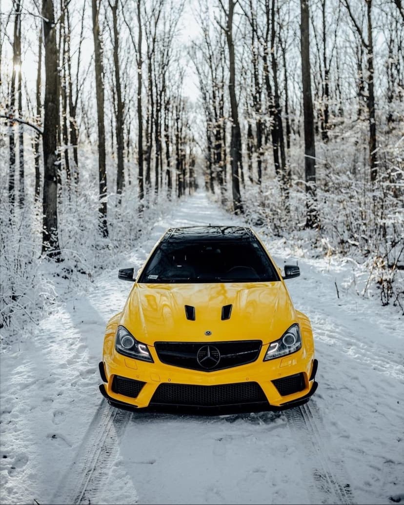 Yellow Mercedes-Benz sports car parked on a snow-covered road surrounded by leafless trees in soft sunlight.