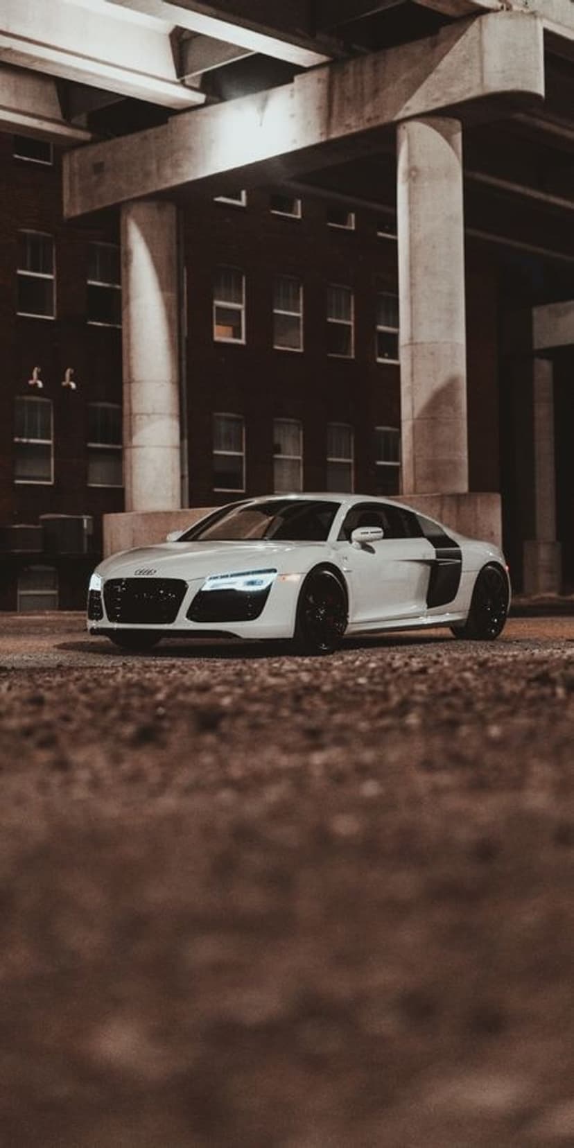 White Audi R8 with glowing headlights parked on gravel under a brutalist concrete bridge at night in the city.