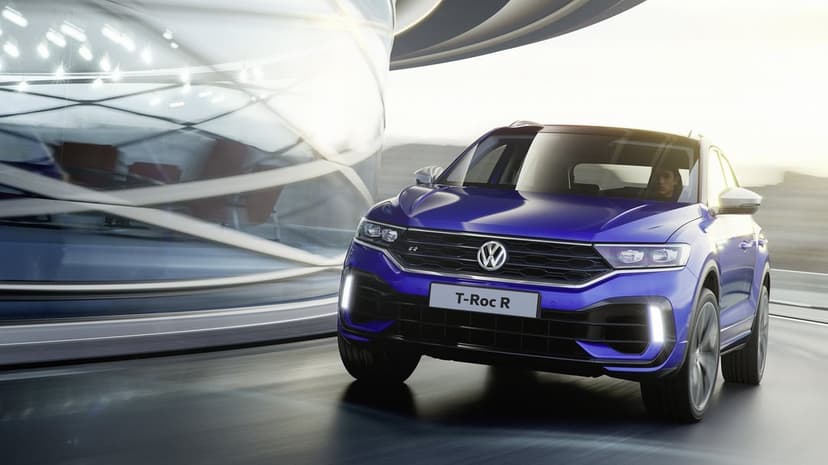 Metallic blue Volkswagen T-Roc R driving past a curved glass building on a motion-blurred road during sunset.