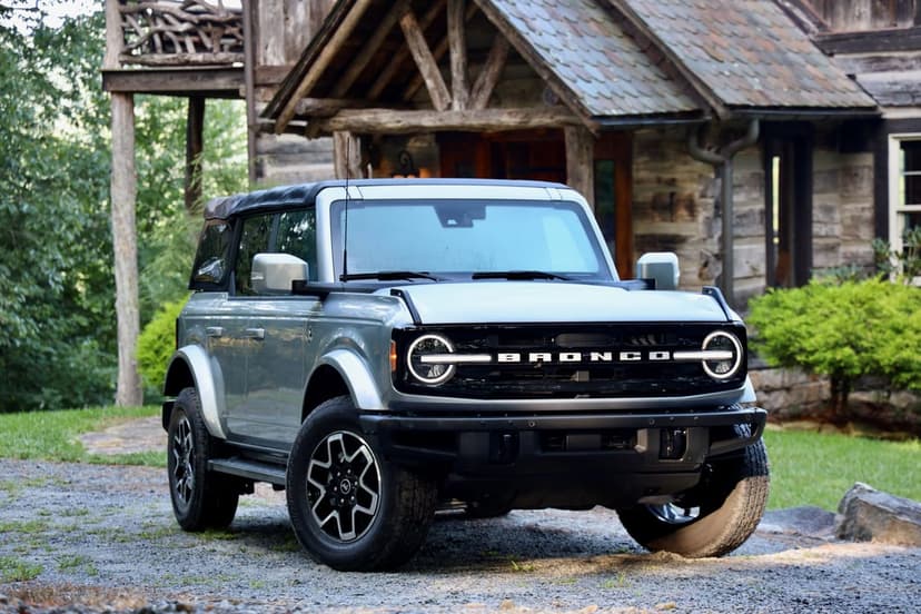 Silver Ford Bronco SUV with round LED headlights parked on a gravel path near a rustic log cabin.