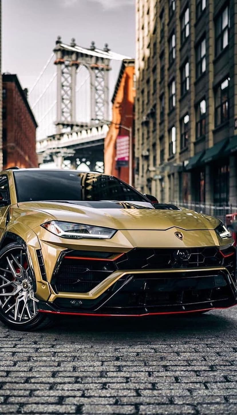 Gold Lamborghini Urus SUV parked on a cobblestone street with the Manhattan Bridge and urban buildings behind it.