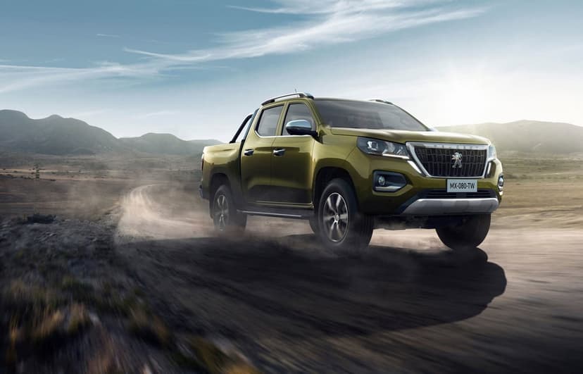 Olive green Peugeot pickup truck driving on a dusty desert road with mountains under a bright sunset sky.