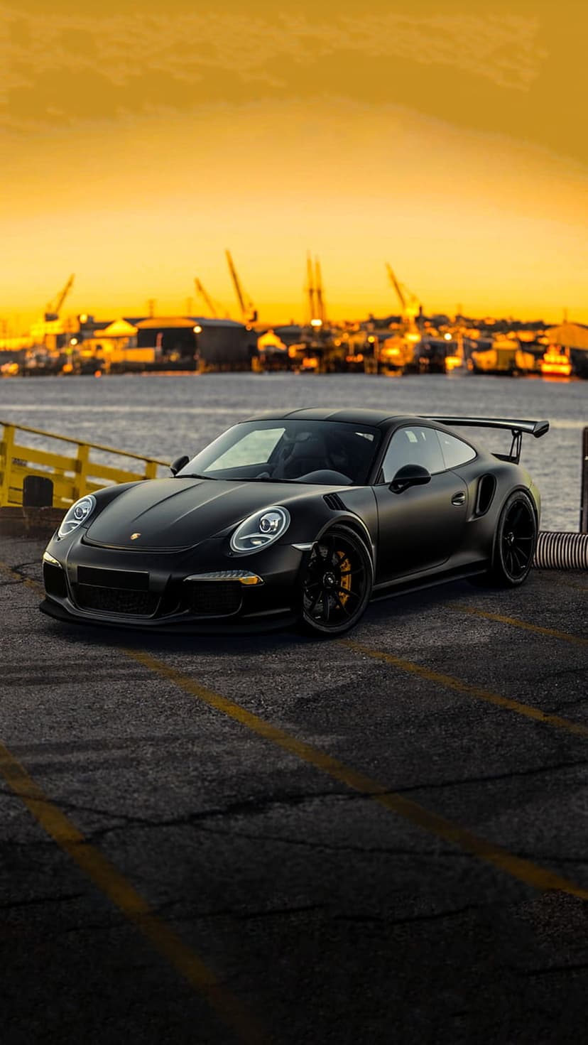 Porsche GT3 RS in matte black parked at an industrial shipyard during sunset with yellow lines on the pavement.