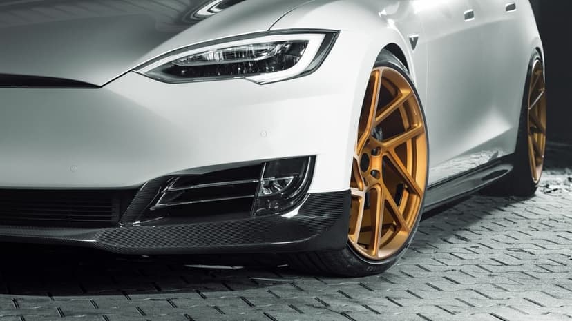 White Tesla Model S with gold concave wheels and carbon fiber splitter parked on a gray cobblestone surface.