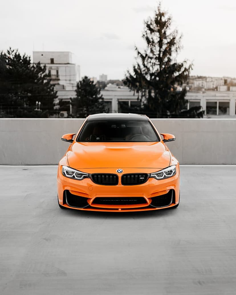 Orange BMW M4 parked on a concrete rooftop featuring a black grille, headlights, and a city building background.
