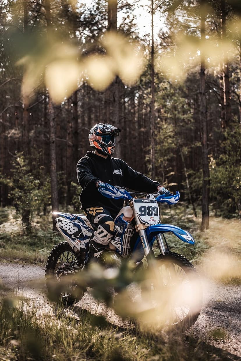 Motocross rider on a blue 989 dirt bike sits on a forest path as sunlight filters through tall pine trees.