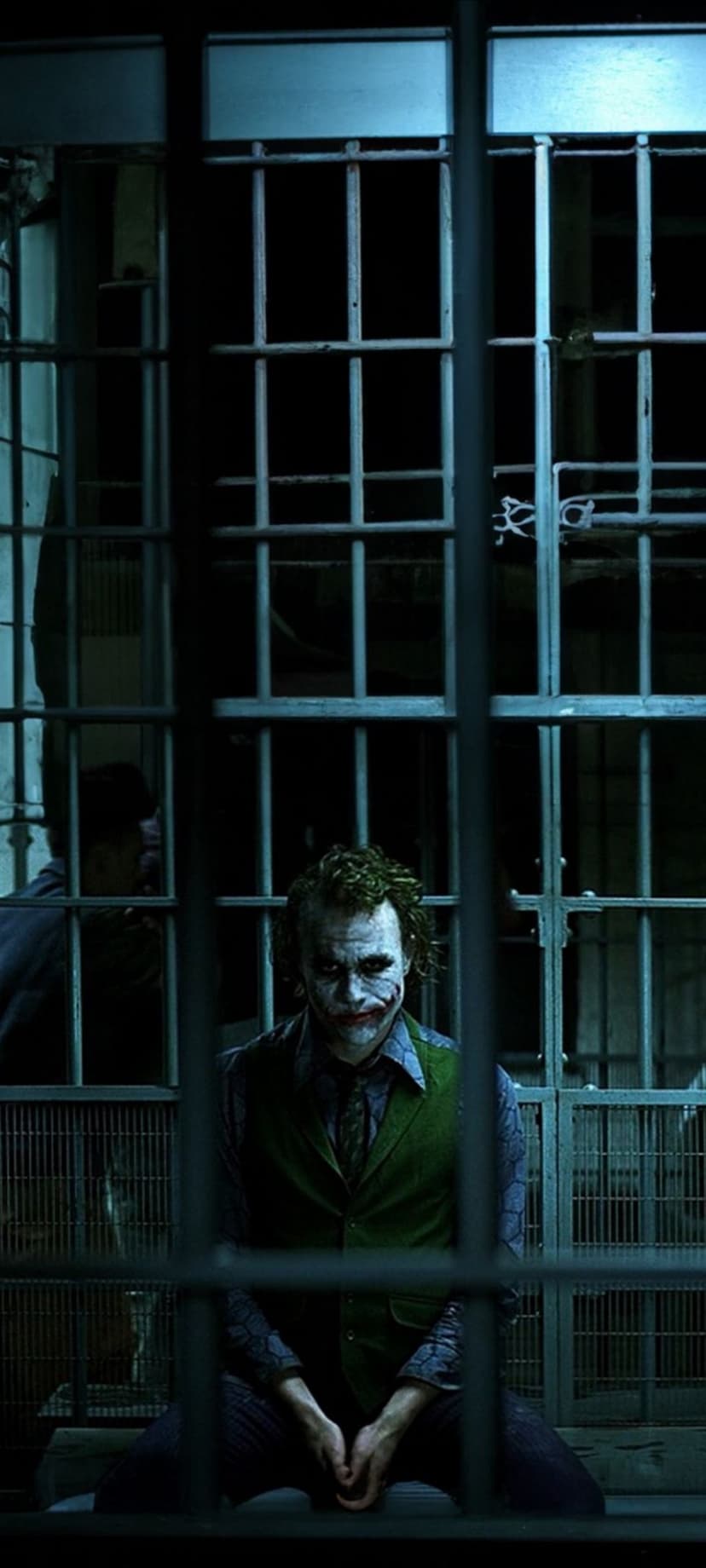 Joker with smudged face paint sits behind metal bars in a dark jail cell with moody shadows and a vacant stare.