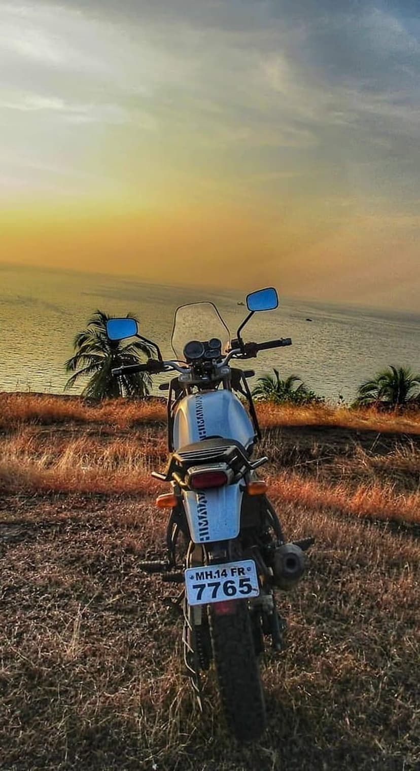 White adventure motorcycle on a grassy cliff overlooking a tropical ocean sunset with orange sky and palm trees.