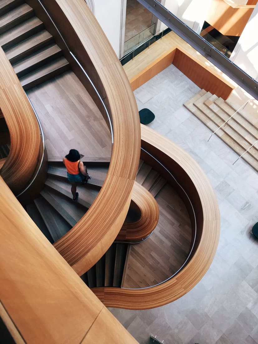 Woman in an orange top walks up a polished wooden spiral staircase under soft natural light in a modern home.