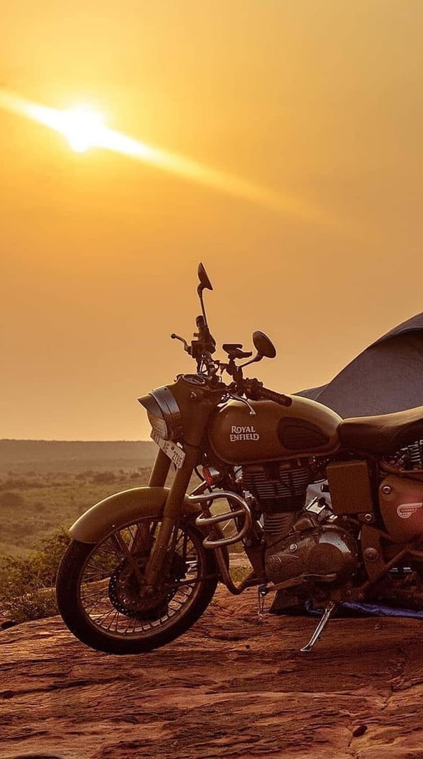 Classic Royal Enfield motorcycle stands on a jagged desert cliff during a warm golden hour with a glowing sun.