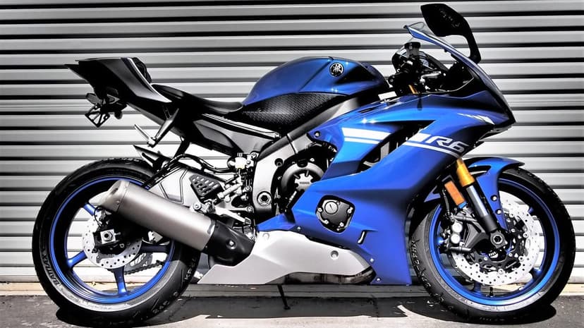 Blue Yamaha R6 sportbike side profile against a gray metal shutter with metallic accents and blue wheels.