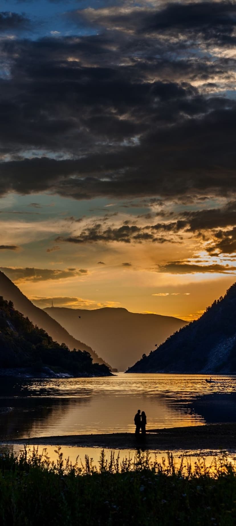 Silhouetted couple on a lakeshore at sunset with dark mountains and orange sky reflections in the water.