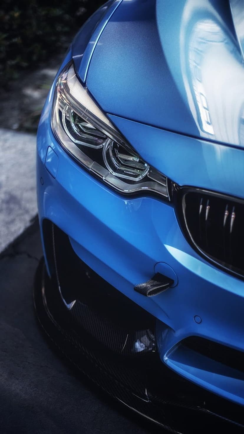 BMW M4 front bumper with metallic blue paint, glowing LED headlights, and dark carbon fiber aerodynamic details.