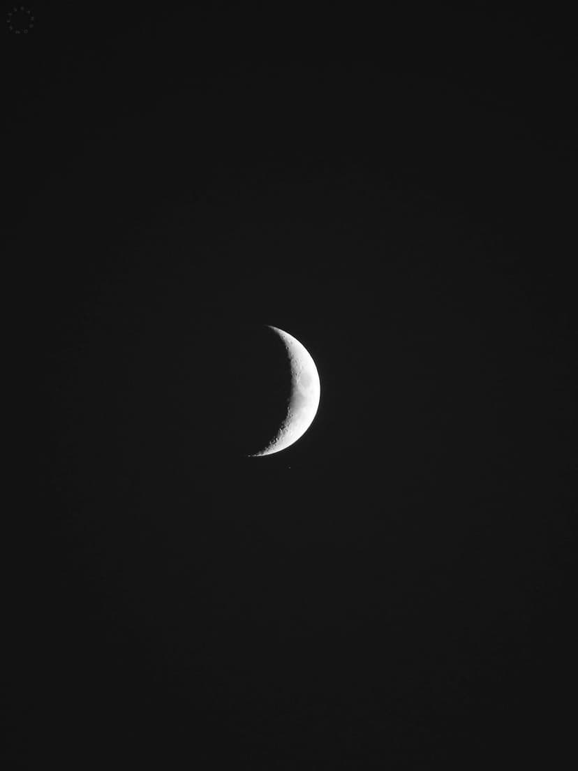 Slender glowing crescent moon showing surface texture against a completely black night sky in portrait view.