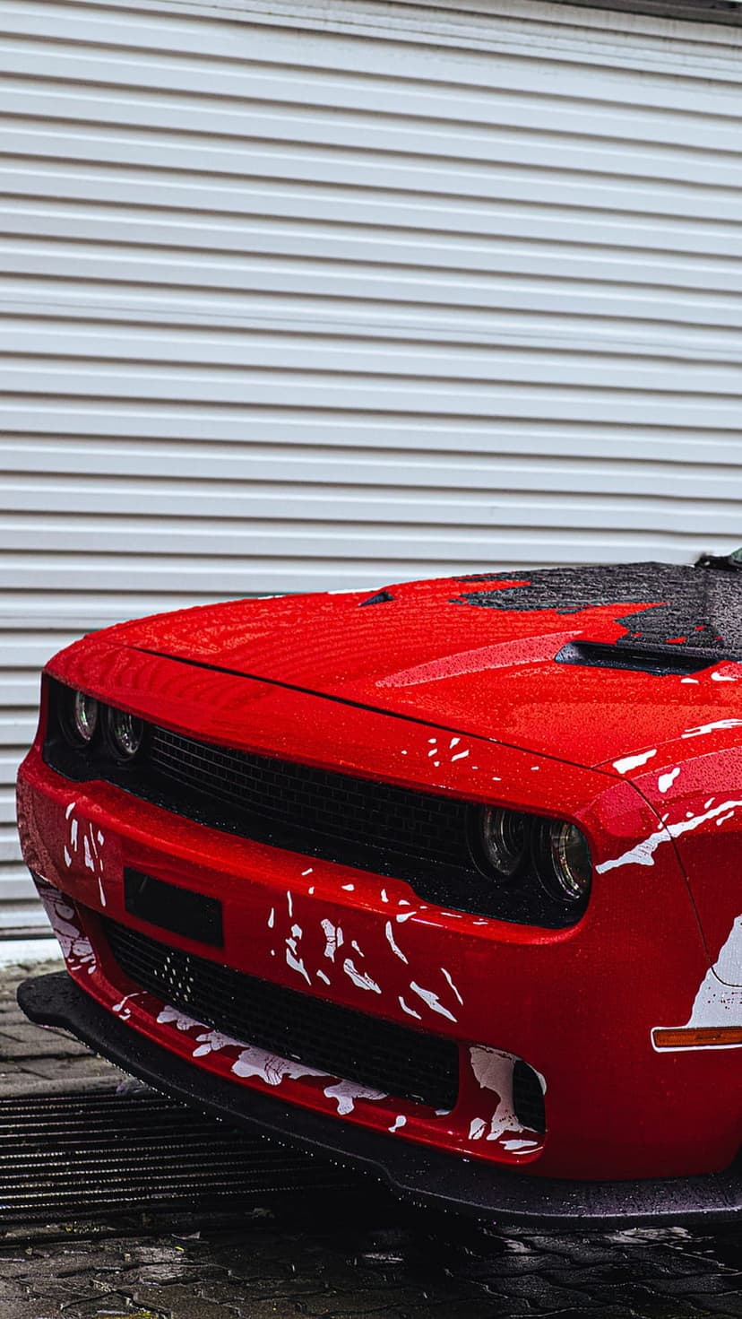 Red Dodge Challenger front grille and headlights with water droplets against a gray ribbed garage door background.