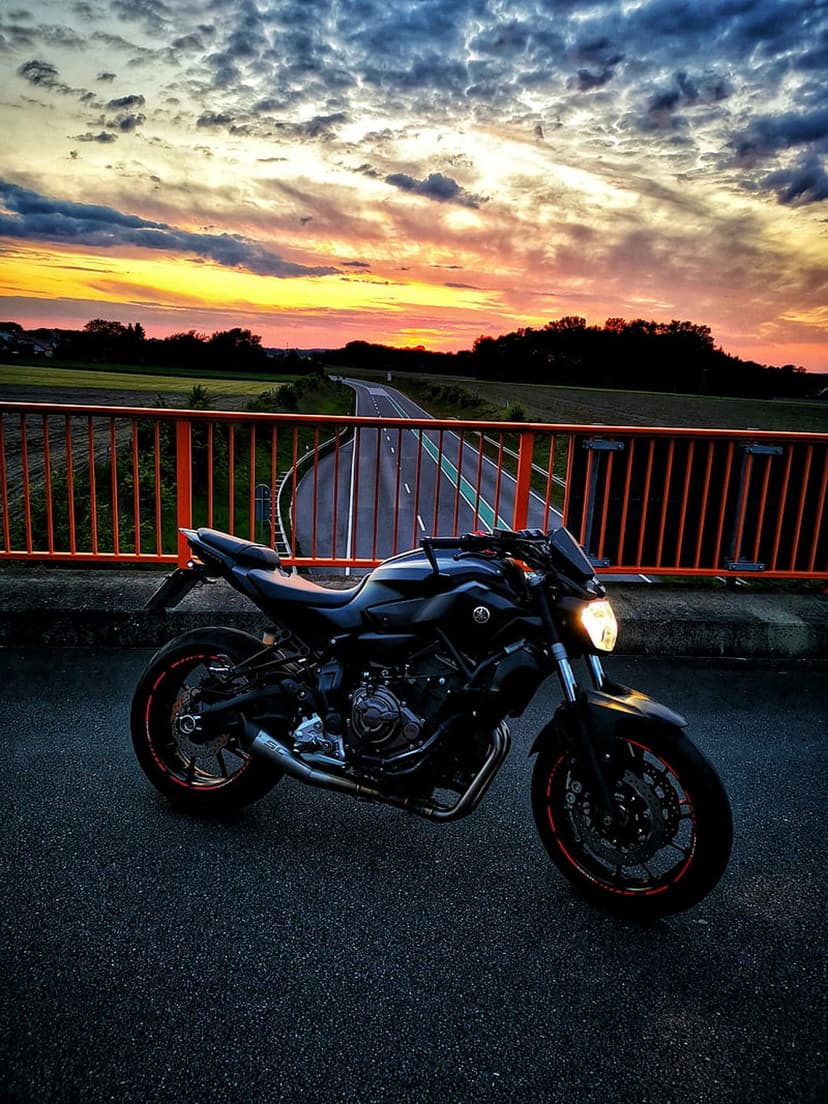 Black motorcycle parked on a bridge with orange railings under a vivid sunset sky of pink and gold clouds.