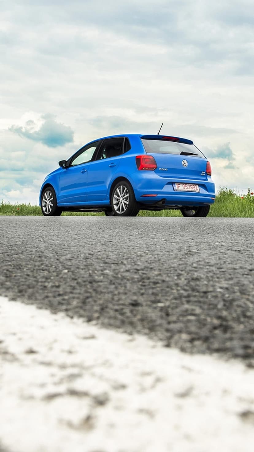 Blue Volkswagen Polo hatchback parked on a paved road under a cloudy sky, shown from a low-angle rear view.