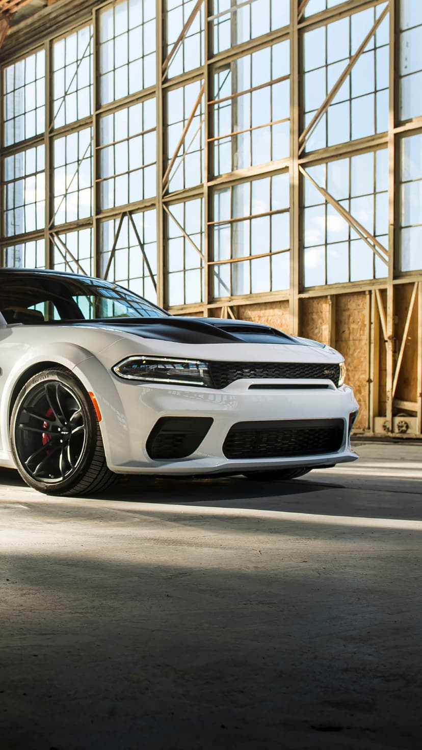 White Dodge Charger SRT Hellcat Widebody with black hood in a sunlit industrial hangar with large windows.