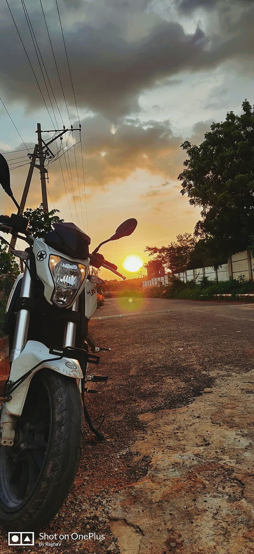 White motorcycle parked on a dirt path under a dramatic sunset sky with heavy clouds and utility poles.