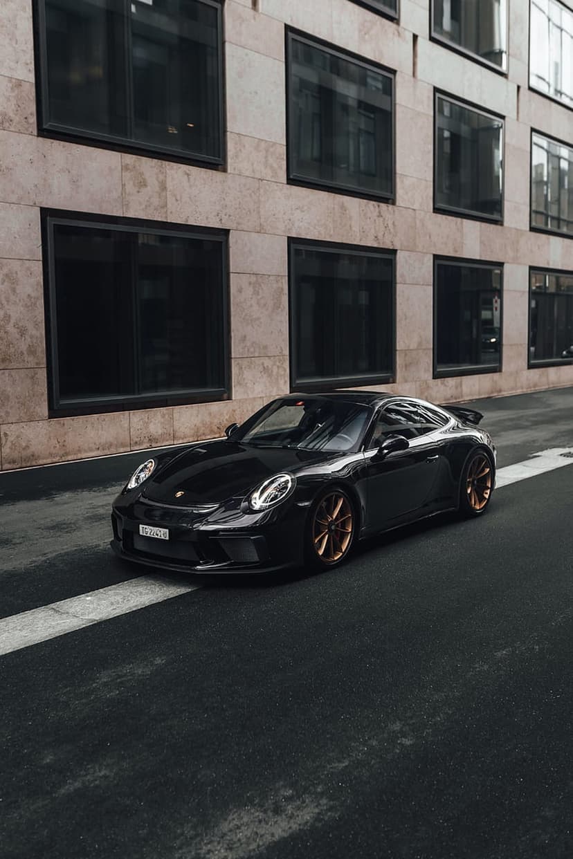 Black Porsche GT3 with bronze wheels parked on asphalt against a glass building with minimalist architecture.