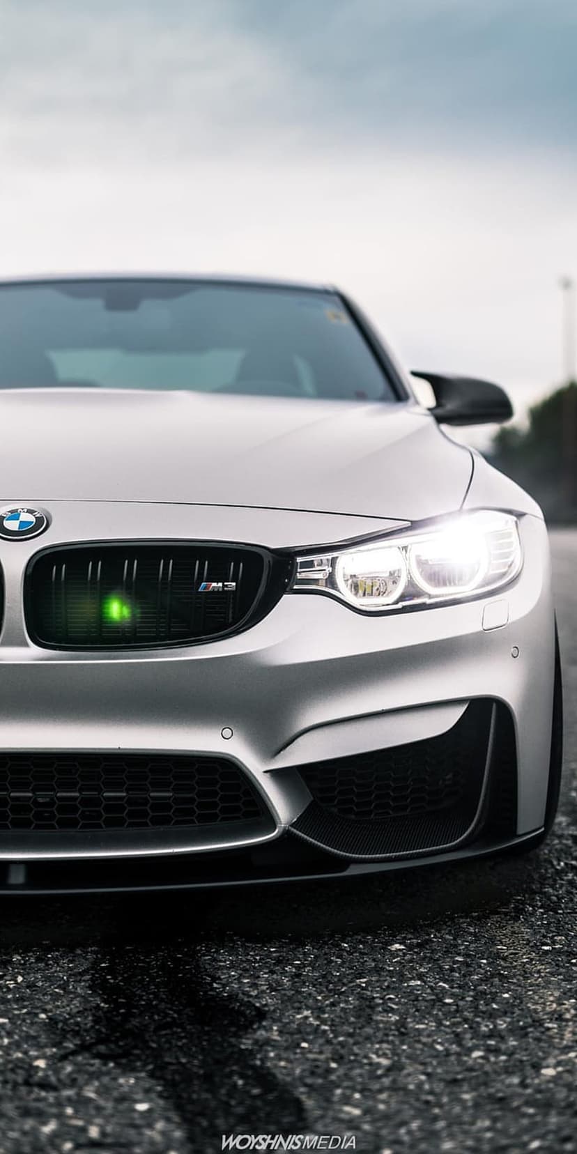 Silver BMW M3 front grille and glowing LED headlights on a damp asphalt road featuring neon green accents.