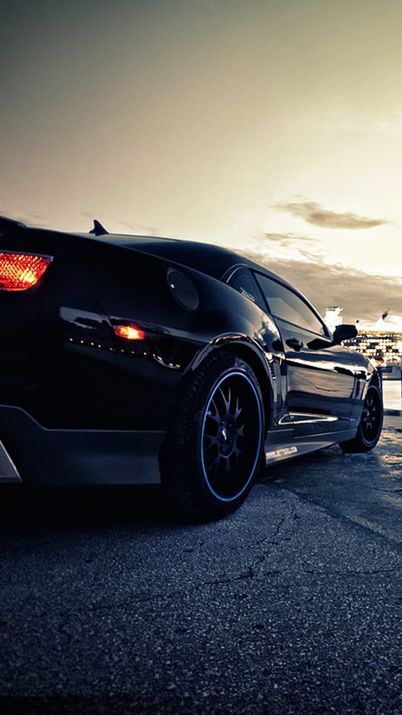 Black muscle car with illuminated red taillights sits on asphalt by water under a cloudy dusk sky at evening.