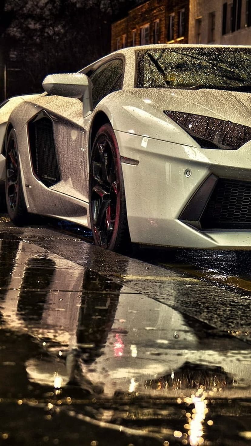 White Lamborghini Aventador parked on wet asphalt at night with streetlights reflecting in surrounding puddles.