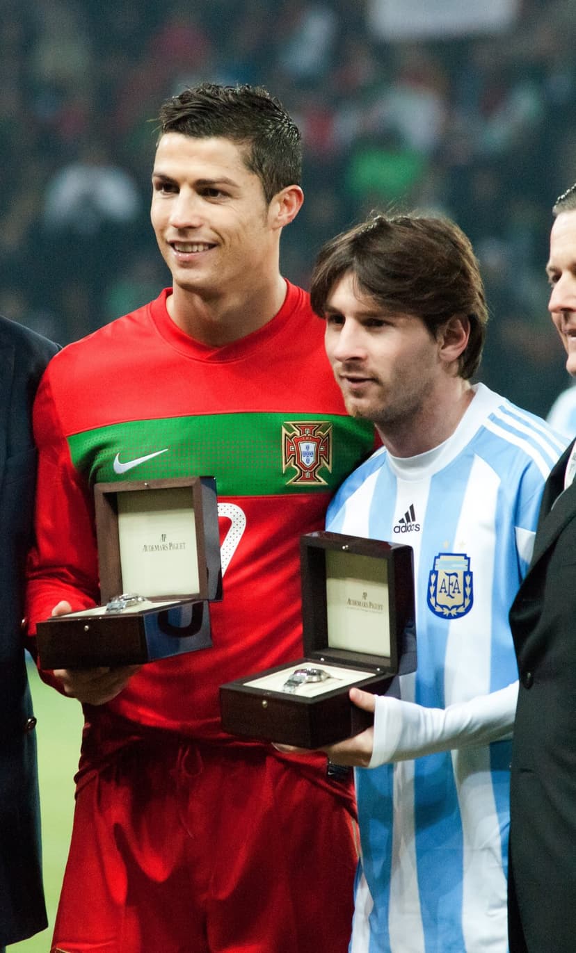 Cristiano Ronaldo and Lionel Messi smiling on a grass pitch while holding wooden boxes with luxury watches inside.