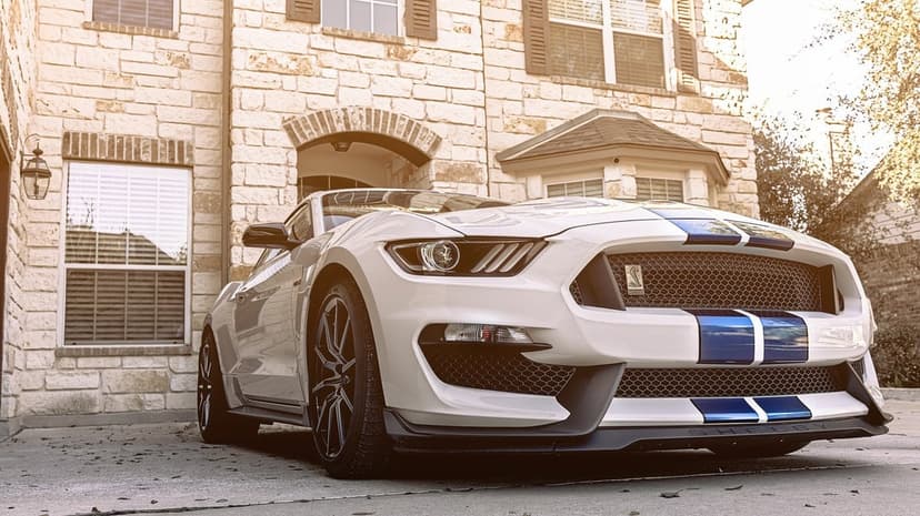White Shelby GT350 Mustang with blue racing stripes parked in front of a stone house under golden sunlight.