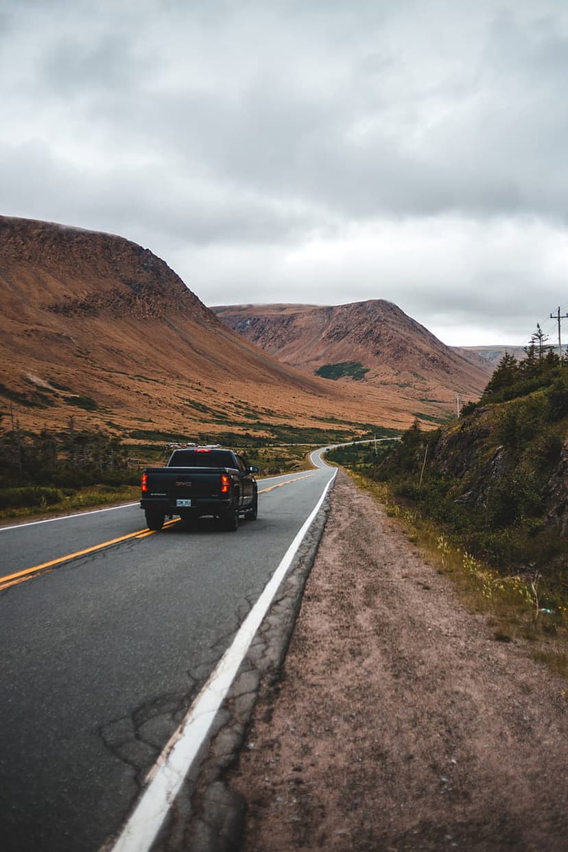 Black pickup truck drives along a winding asphalt road between golden mountain slopes under a cloudy gray sky.