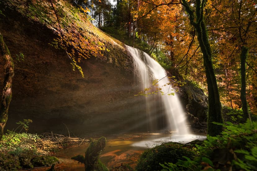 Waterfall cascading over mossy rocks amidst sunlit orange autumn trees and rising mist in a vertical forest view.