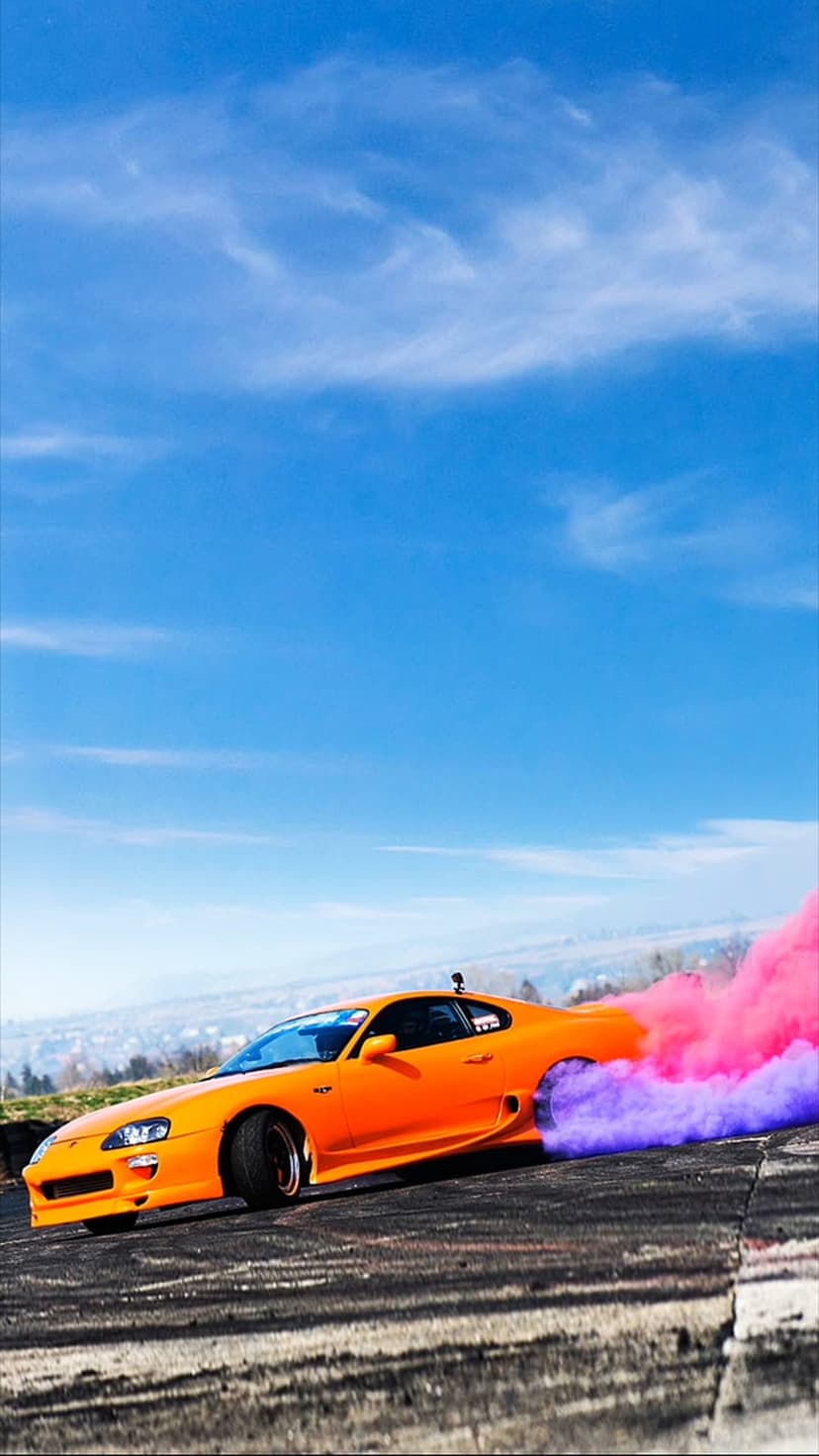 Orange Toyota Supra drifting on a track with thick pink and purple smoke clouds under a bright blue sky.