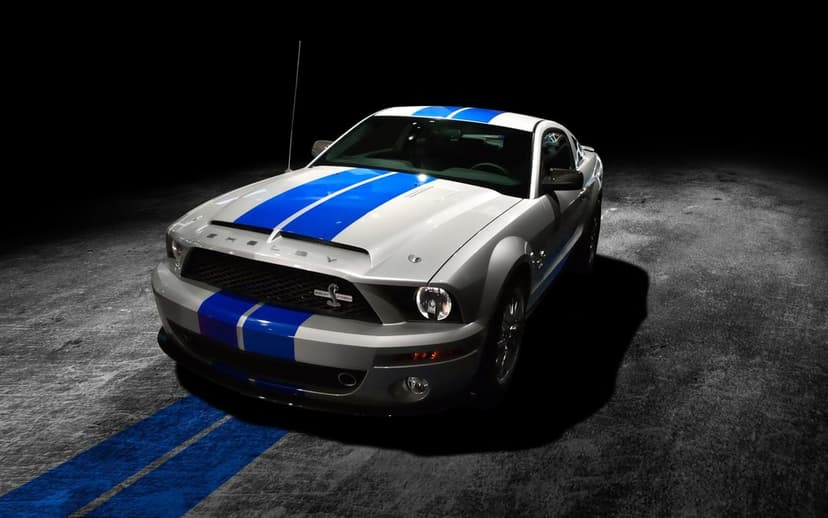 Silver Shelby GT500KR muscle car with dual blue racing stripes parked on textured ground in a dark spotlight.