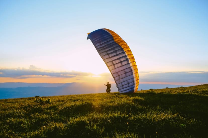 Paraglider on a grassy hill prepares to launch with an inflated wing silhouetted against a golden sunset sky.