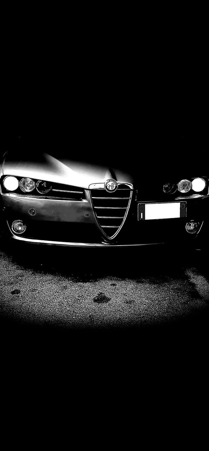 Alfa Romeo car front featuring a triangular grille and lit headlights on a dark asphalt road in black and white.