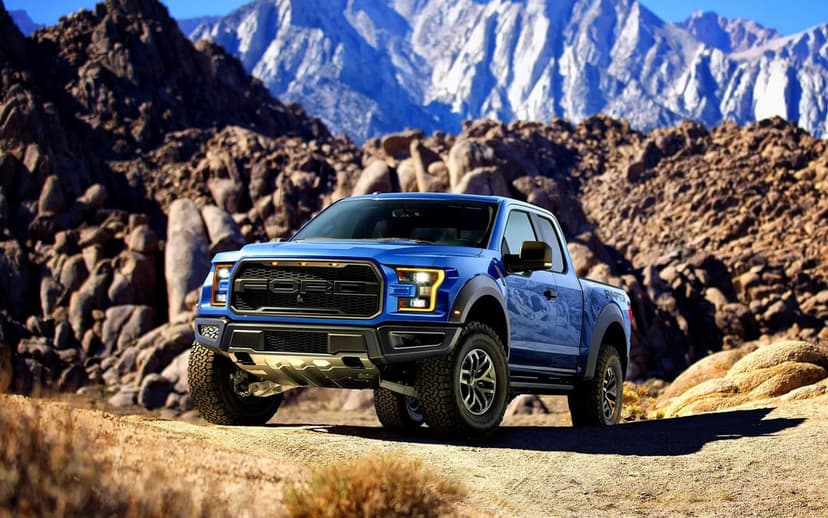 Blue Ford Raptor pickup truck parked on a rocky desert trail with snow-capped mountains in the distance.