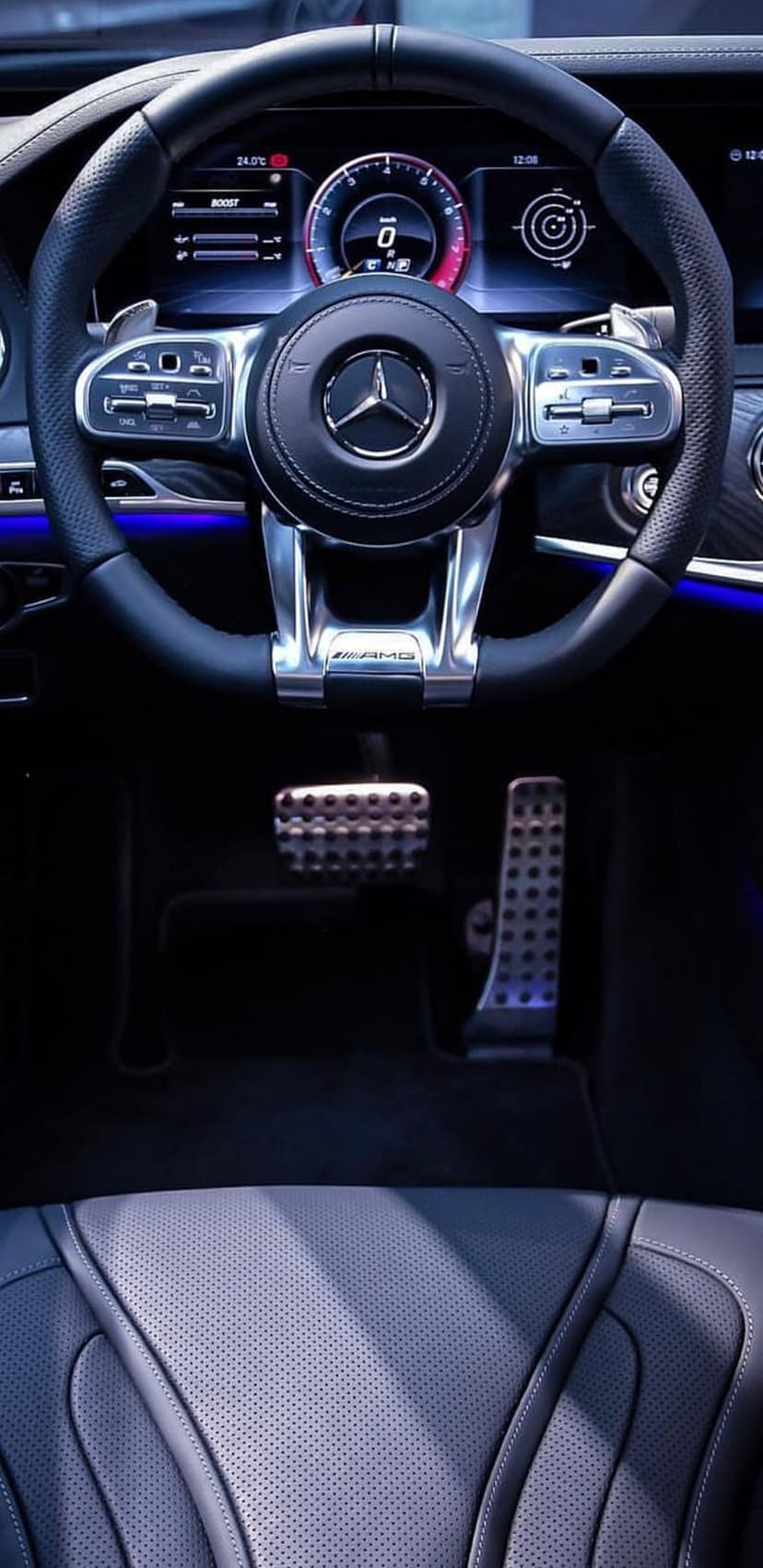 Mercedes-Benz AMG steering wheel and brushed metal pedals illuminated by blue ambient light in a luxury car cabin.