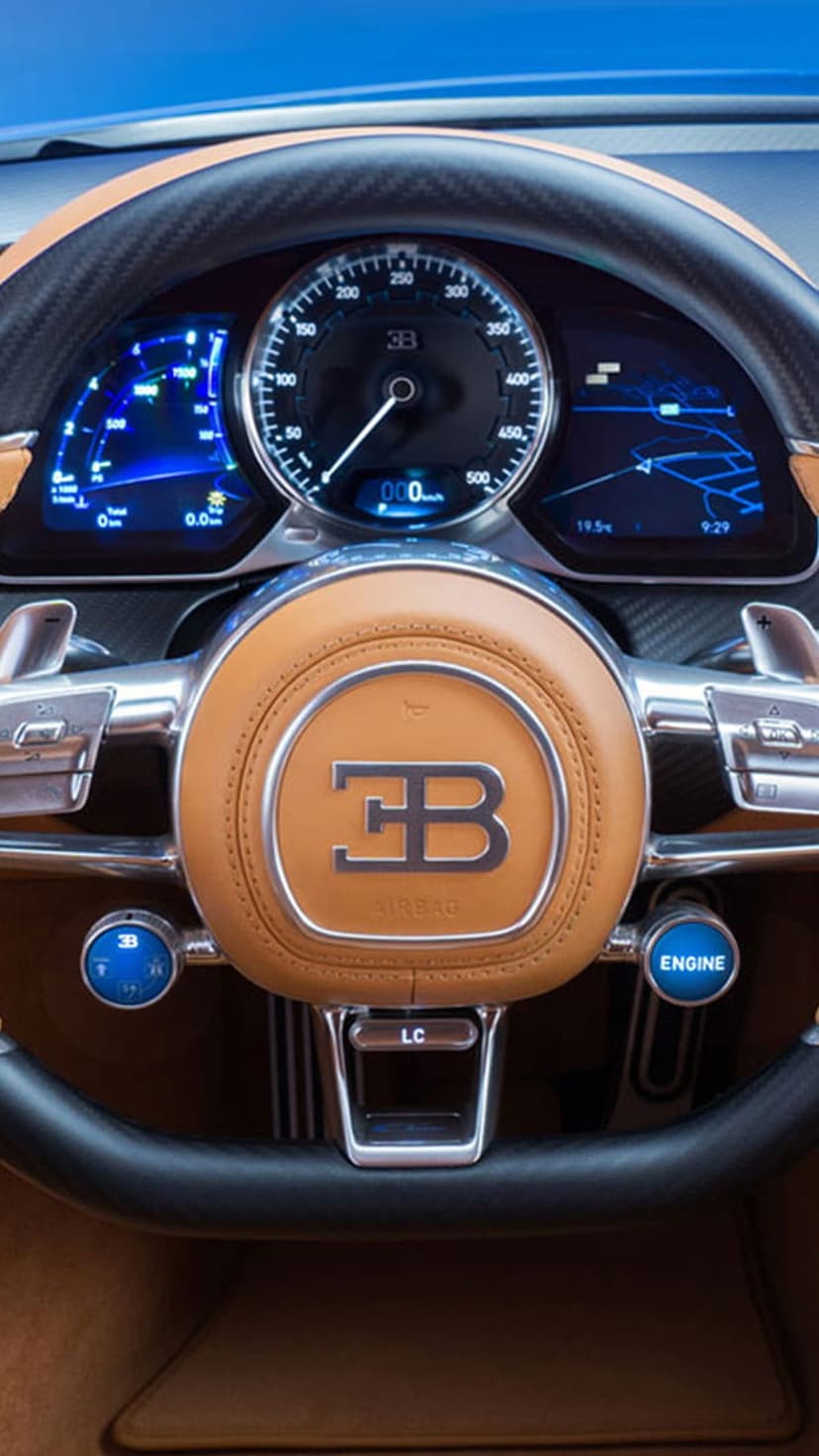 Bugatti steering wheel with a glowing emblem and digital dash displays set in a carbon fiber hypercar interior.
