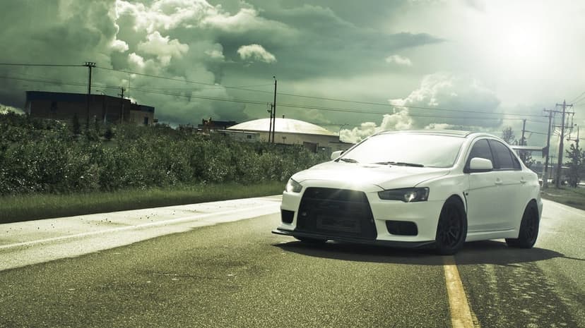 White Mitsubishi Evolution X sports car on a road under a moody, overcast sky with industrial buildings.