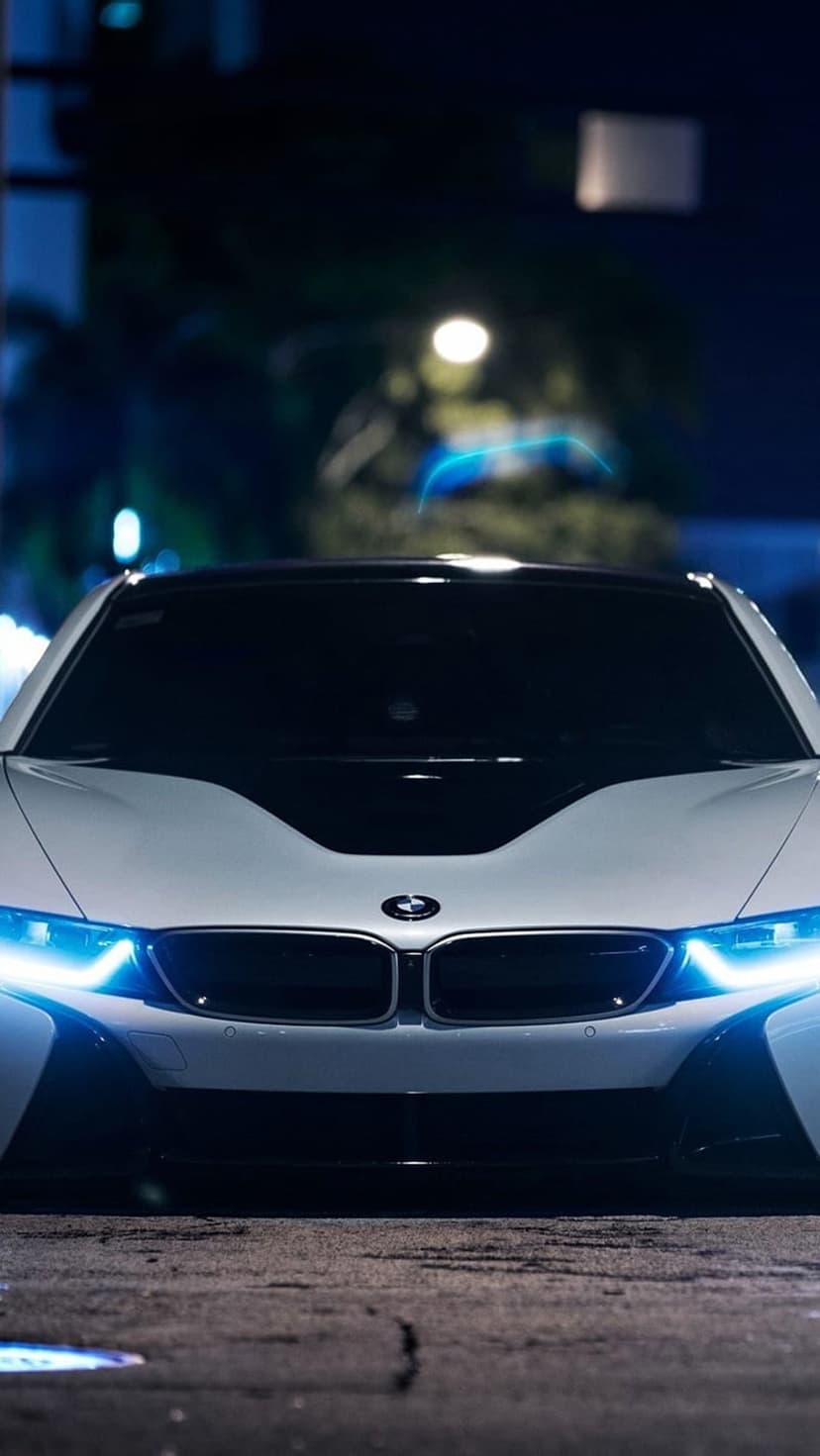 BMW i8 front fascia with glowing blue headlights and a white hood parked on a dark asphalt street at night.