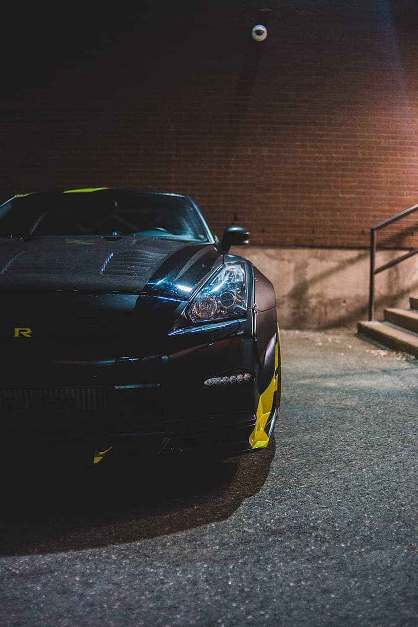 Black sports car with yellow trim and carbon fiber hood parked on a dark city street near brick stairs at night.