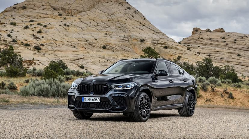 Black BMW X6 M Competition parked on gravel in front of large layered rock formations under a cloudy sky.