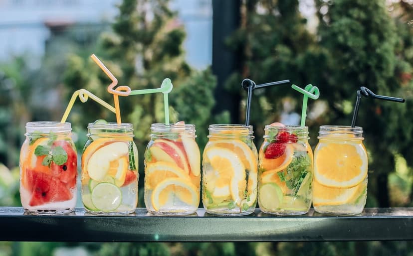 Six mason jars containing citrus, berries, and mint in water with colorful straws on a bright summer background.