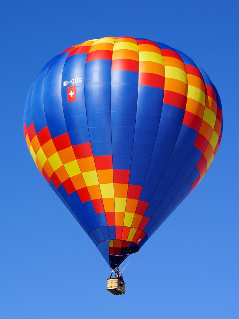 Swiss hot air balloon with red, yellow, and blue panels soaring in a cloudless azure sky during a daytime flight.