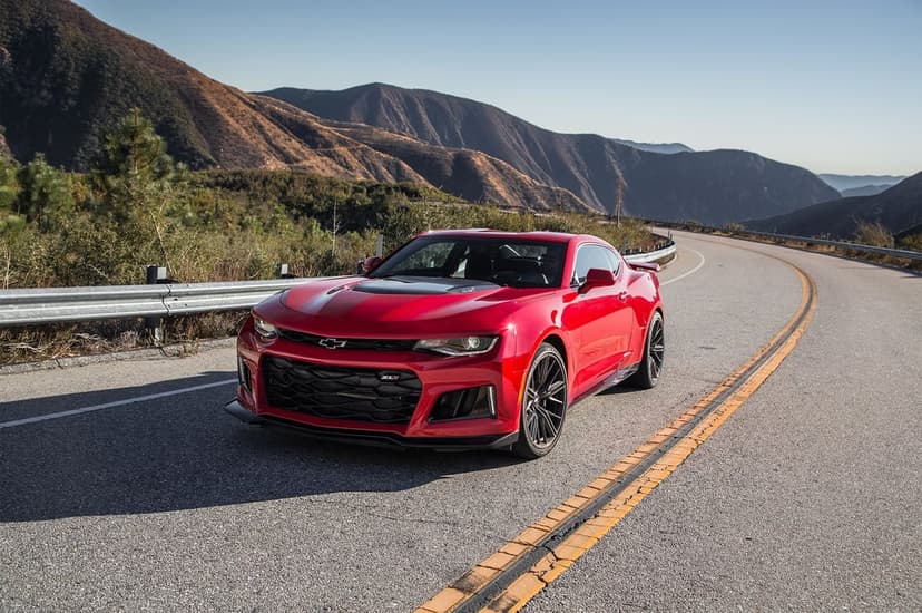 Red Chevrolet Camaro ZL1 driving on a winding mountain road under a clear blue sky with hills in the background.